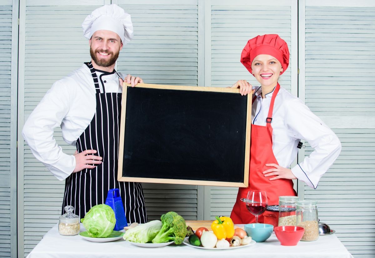 Making good food. Chef and cook helper teaching master class. Couple of man and woman holding empty blackboard in cooking school. Master cook and prep cook giving cooking class, copy space.