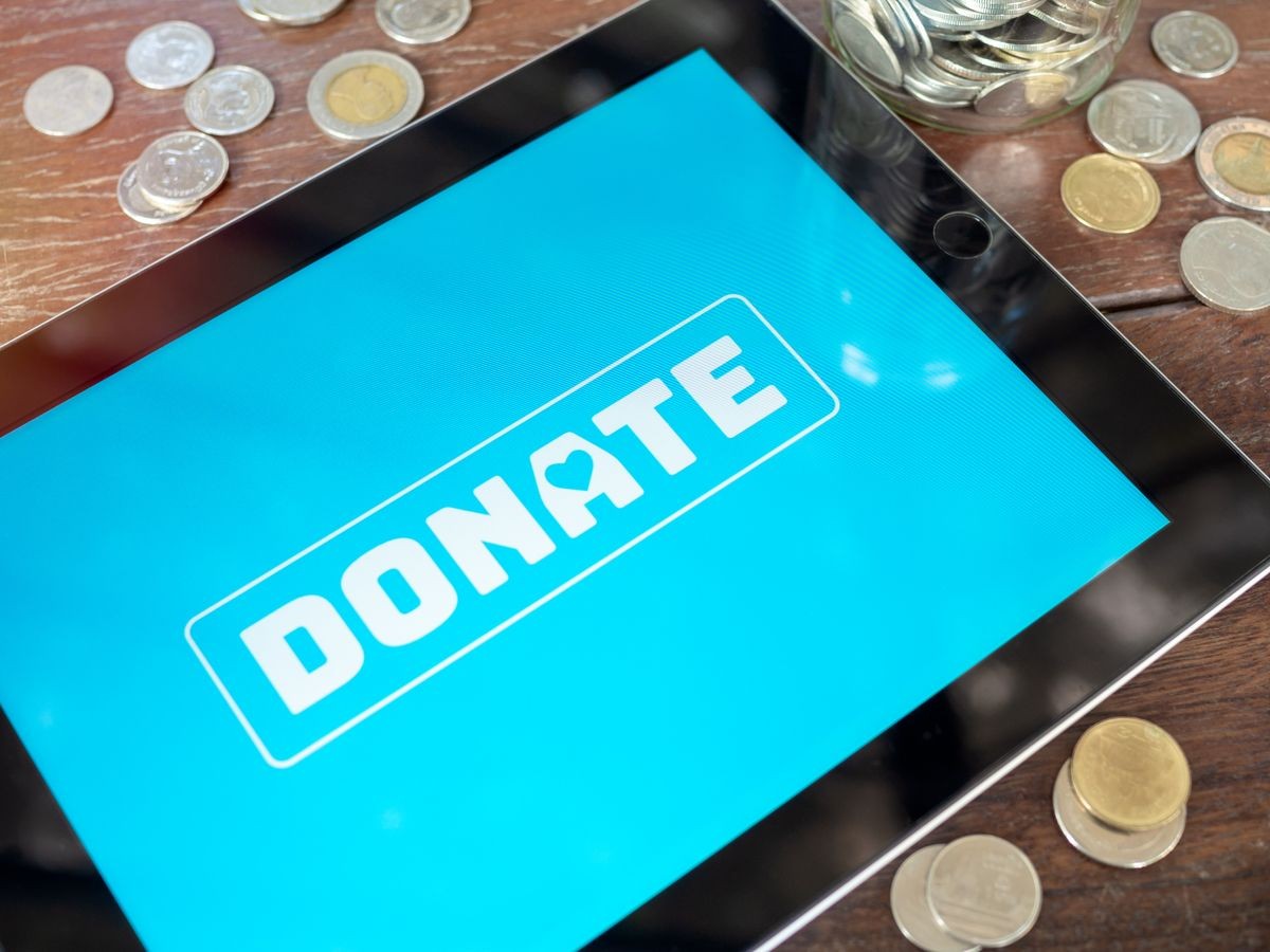 Close-up donate icon button on blue screen on tablet with coins on wooden table. Donation online concept.